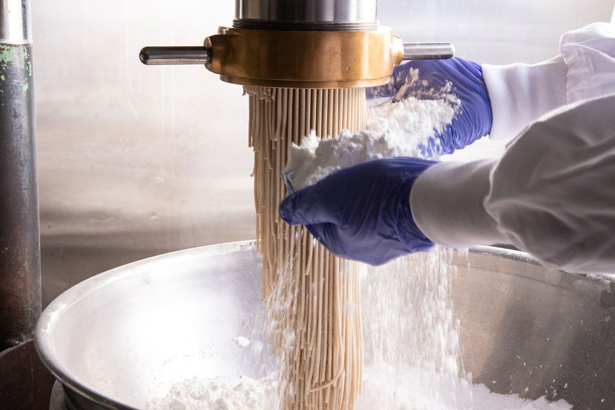 「芳華冷麺」の特製麺が製麺機から出てきた直後、職人が丁寧に打ち粉をまぶしている製造工程の画像。コシと風味を生み出す、こだわりの麺作り。