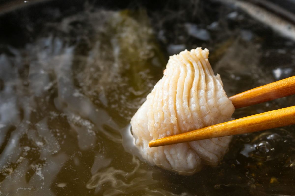 大分県産の鱧の切り身を鍋の出汁にくぐらせ、しゃぶしゃぶしているアップ画像。繊細で上品な旨味を持つ鱧の身が、花のように開く様子。