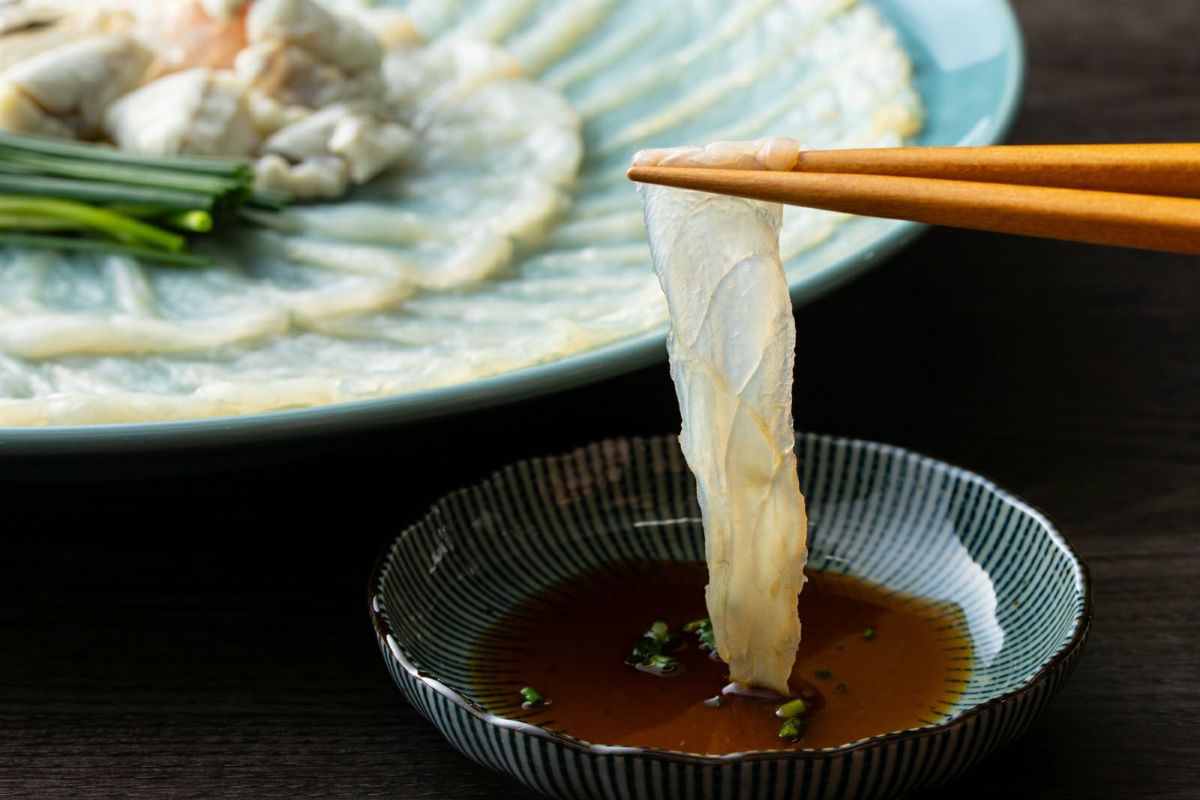 豊後とらふぐの刺身を、付属の特製ポン酢につけているアップ画像。ふぐ刺しの美しい身の締まりと、薬味の風味が伝わる、大分からのお取り寄せ品。