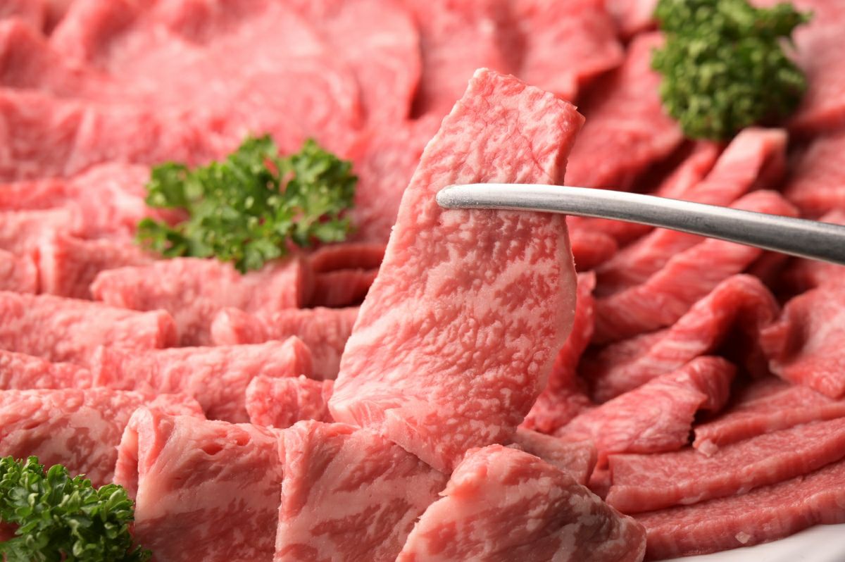 大分県のブランド牛「おおいた和牛」のカルビとロースをきれいに盛り付けた皿を背景に、霜降りの美しい一切れをトングで持ち上げているアップ画像。焼肉に最適なセットを通販でお取り寄せ。