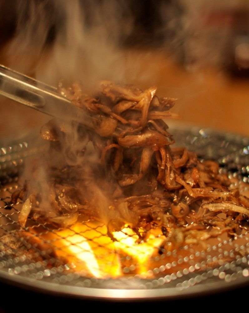鶏皮を炭火でじっくりと焼いている調理中の画像。炎と煙の中で香ばしい焼き色がつき、特製の甘辛いタレが絡みつく様子。